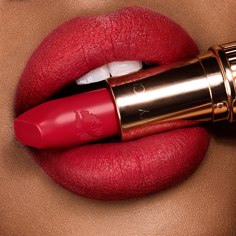 Lips close-up of medium-tone model wearing a warm-red matte lipstick and holding the lipstick in her mouth. 