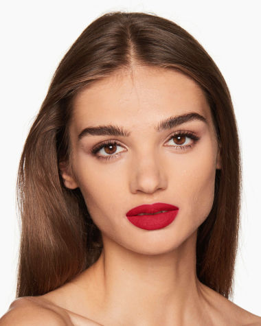 A medium-tone model with brown eyes wearing a matte, bright, cherry-red lipstick.