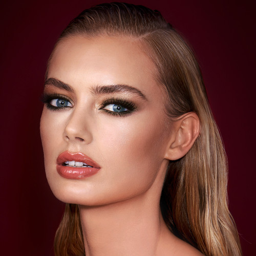A light-tone model with blue eyes wearing smokey green eye makeup with black eyeliner, soft brown blush, and orange-red lipstick with gloss on top. 