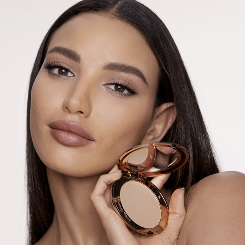 Medium-tone brunette model with flawless, matte skin wearing a nude pink lipstick and holding an open, pressed powder compact in a medium shade.