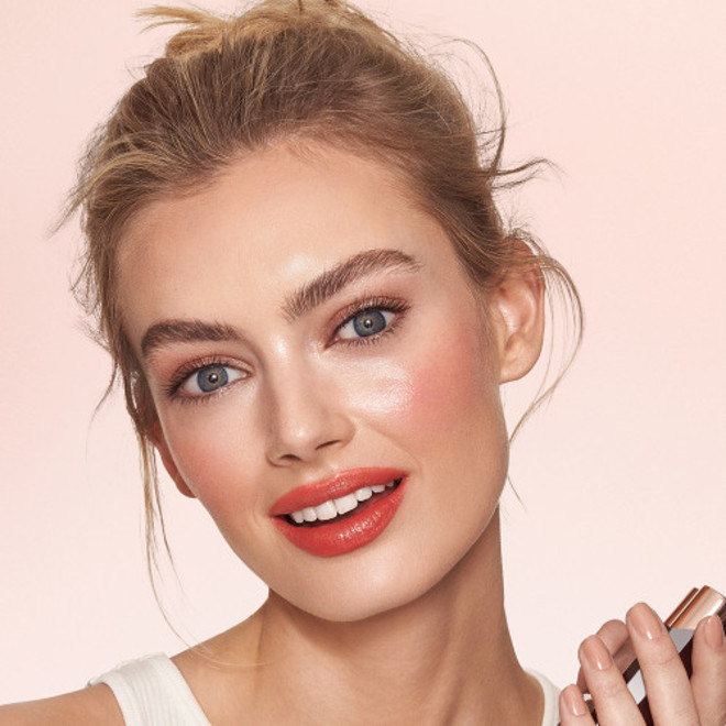 Fair-tone model with blue eyes wearing shimmery champagne pink eyeshadow with vivid coral blush and lip stain, and dewy opal-coloured highlighter for a fresh, glowing makeup look.