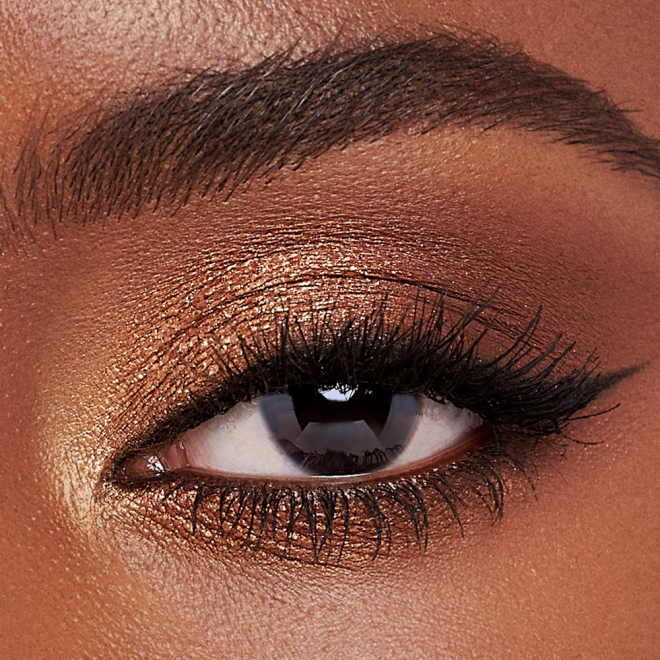 Single-eye close-up of a deep-tone model with brown eyes wearing metallic honey gold, soft amber, and dark chocolate brown eye makeup. 