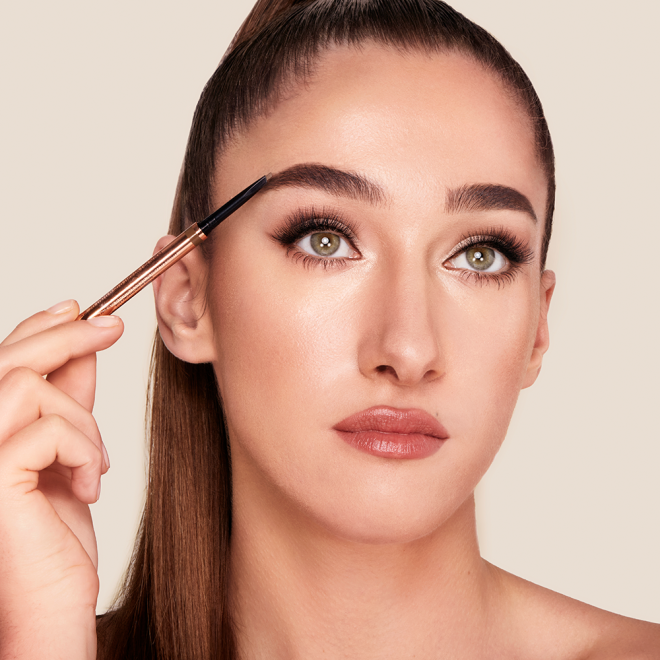 Light-tone model with green eyes filling in her brows with a brown-coloured eyebrow pencil.