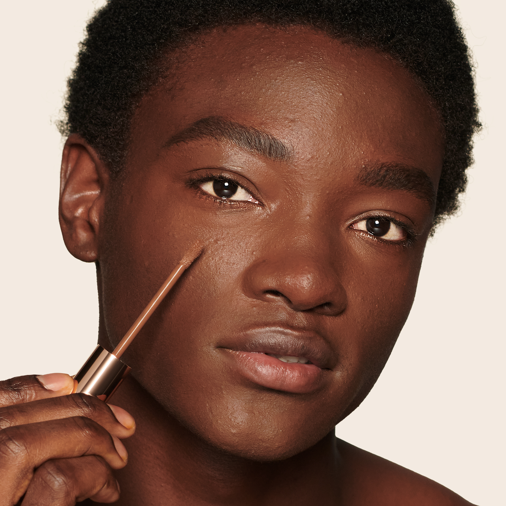 Deep-tone model with brown eyes applying a radiant, concealer that brightens, covers blemishes, and makes her skin look fresh and wearing nude lip gloss and subtle eye makeup.