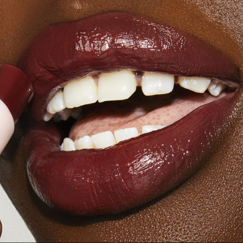 Lips close-up of a deep-tone model applying a moisturising lipstick balm in a berry shade with a high-shine finish.