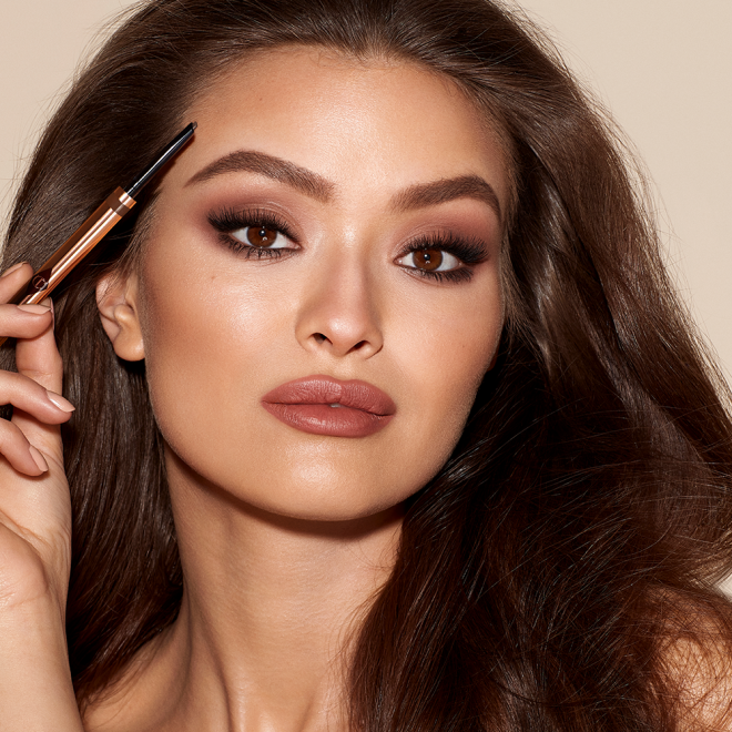 A medium-tone model with brown eyes wearing smokey brown eye makeup with a muted brick-red lipstick and applying a medium-brown-coloured brow gel.