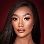 A deep-tone model with brown eyes wearing shimmery copper and gold eye makeup with black eyeliner, glowy bronzed cheeks, and pinkish-brown lipstick with gloss on top. 
