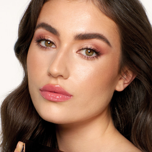 Light-tone brunette model with hazel eyes wearing shimmery rose gold and champagne eyeshadow, glowy pinky-peach blush, soft gold highlighter, and glossy lipstick in a rosebud-pink colour.