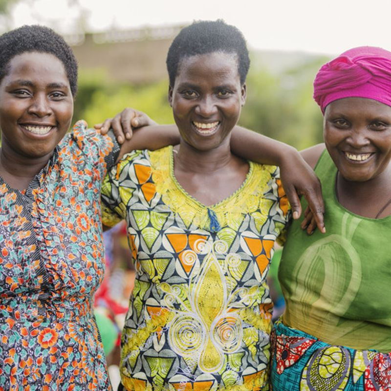 Three deep-tone ladies wearing bright-colored dresses and smiling at the camera. 