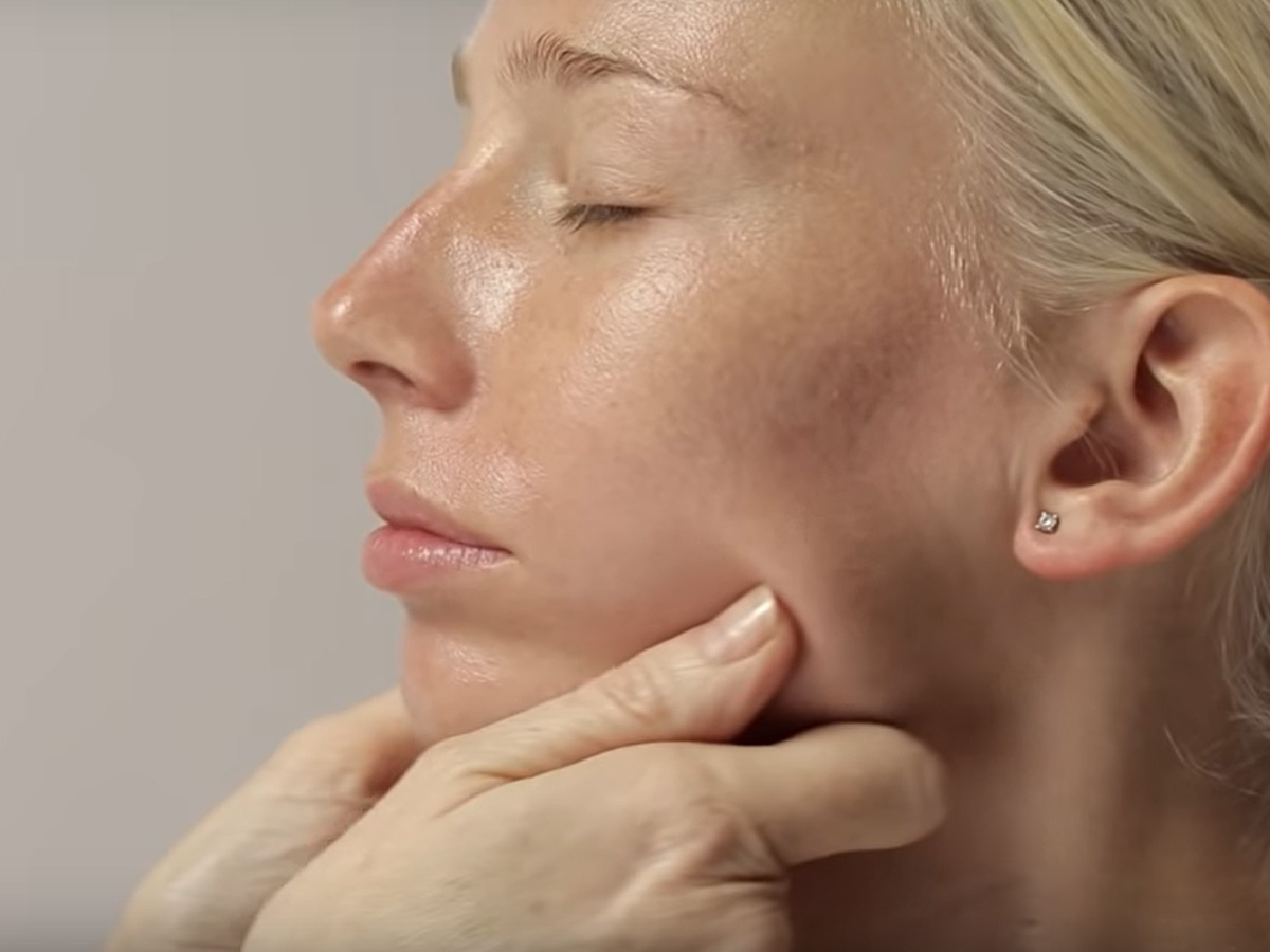 A light tone, blonde model getting a dewy, face cream with a luminous finish applied on her jawline.