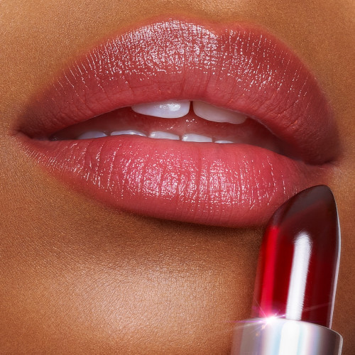 Lips close-up of a deep-tone model wearing a subtle nude red lipstick while holding a glowy, crystal-like red lipstick against her lips. 