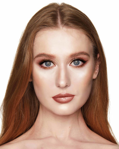 A fair-tone model with blue eyes wearing shimmery copper and gold eye makeup with black eyeliner, glowy bronzed cheeks, and pinkish-brown lipstick with gloss on top. 