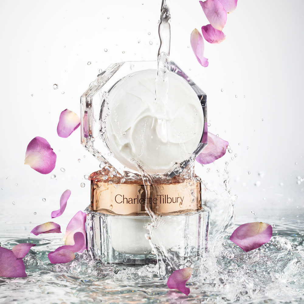 Two pearly-white face creams, one with a gold-coloured lid and the other without, in glass jars with sparkling water and rose petals falling on them. 