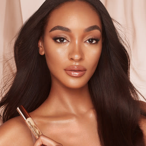 Deep-tone model with glowy, radiant, and flawless skin, wearing nude pink lip gloss with taupe eyeshadow, and black eyeliner while holding a concealer.