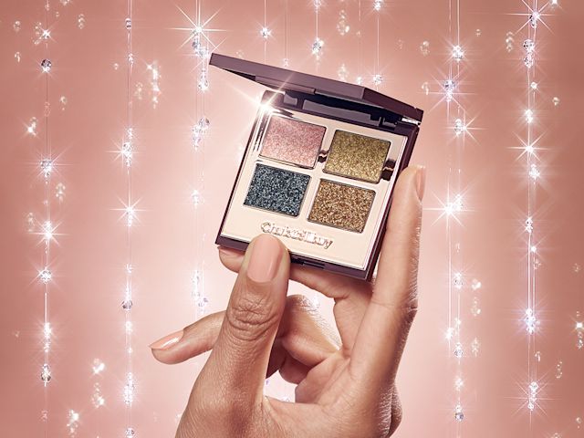 Close-up of a medium-tone model's hands holding an open, mirrored-lid quad eyeshadow palette with shimmery eyeshadows in rose gold, teal, honey-gold, and bright gold colours. 