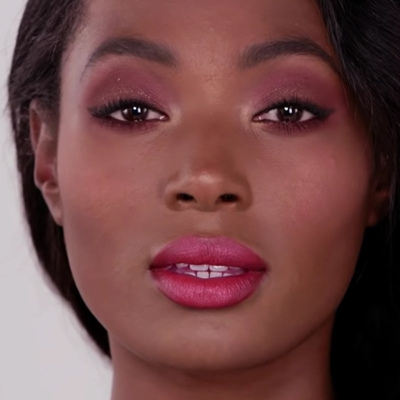 Close-up of a deep-tone model with brown eyes wearing smokey plum and red eye makeup with bright pink lips. 