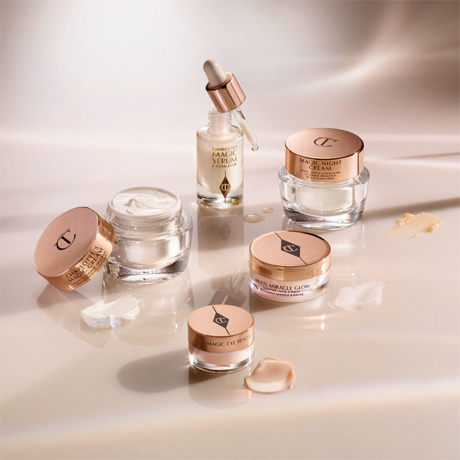 Banner with a cleansing balm in a glass jar, facial serum in a glass bottle and its dropper lid next to it, eye cream, day, and night creams in glass jars with gold-coloured lids. 