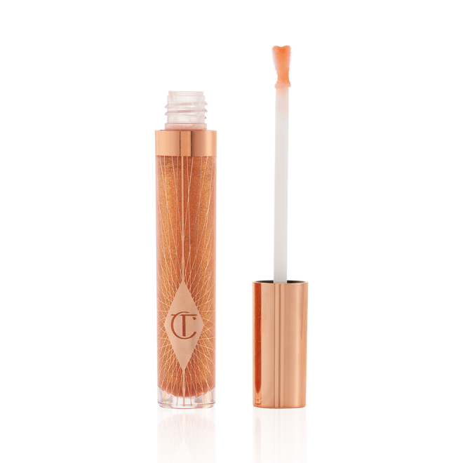 A lip gloss in a sheer gold shade in a glass tube with its heart-shaped applicator next to it.