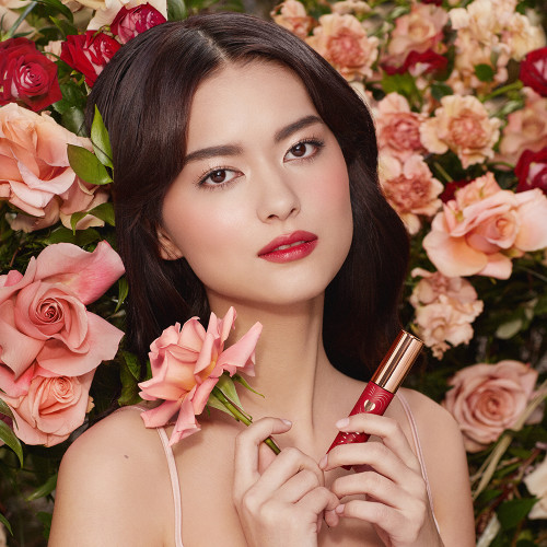 A fair-tone brunette model with brown eyes wearing a vibrant, poppy-red lip tint and holding the lip tint in one hand and a flower in the other.