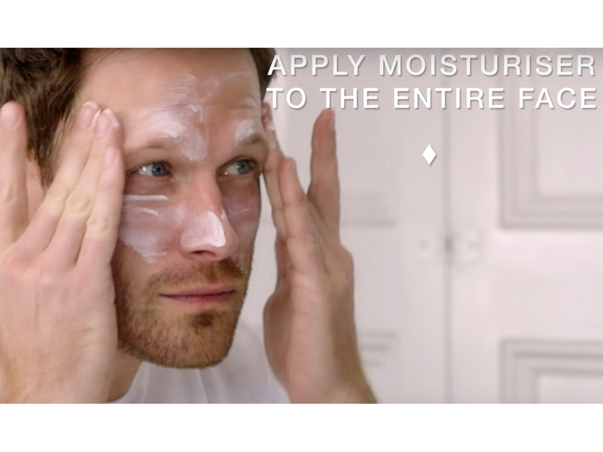 A light-tone, brunette male model patting a face cream on his forehead. 