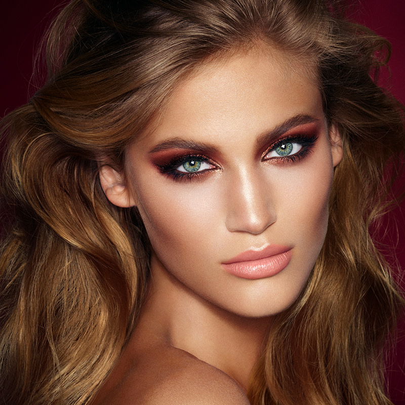 A light-tone model with blue eyes wearing smokey brown and gold eye makeup with warm pink blush and glossy nude-pink lips