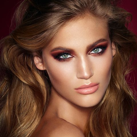 A light-tone model with blue eyes wearing smokey brown and gold eye makeup with warm pink blush and glossy nude-pink lips