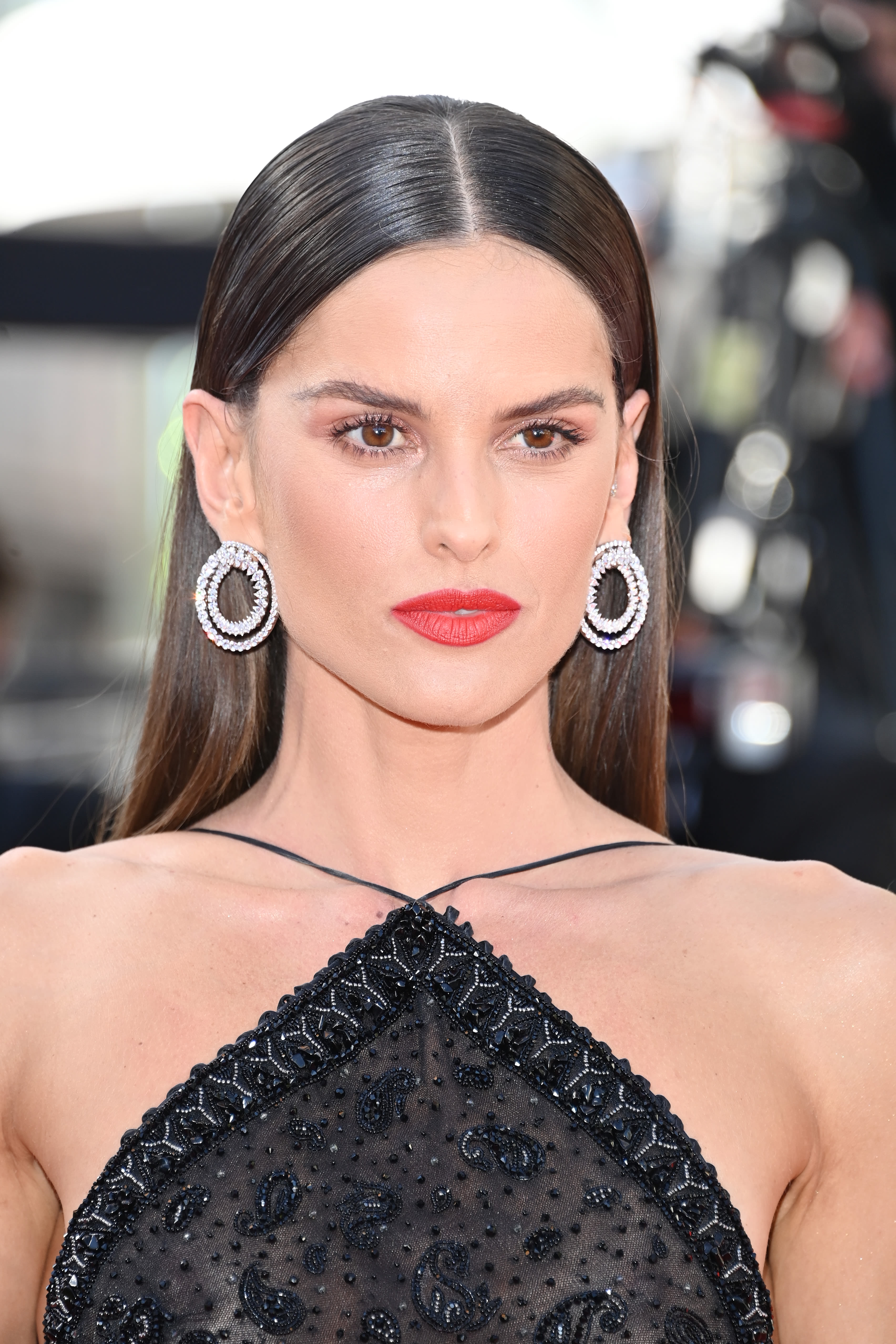 Iza Goulart at Cannes Film Festival 2021, wearing smokey eye makeup with a bold red lipstick and a black dress with silver, dangly earrings.