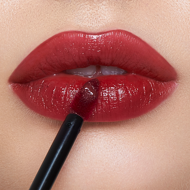 Tinted Love in Tripping on Love on light-tone model lips close-up Lips close-up of a light-tone model applying a vibrant, cherry-red lip tint with a doe-foot brush applicator.