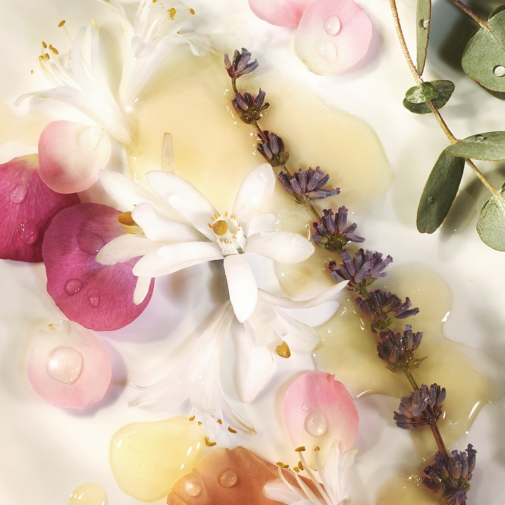 Banner with different flowers, flower petals, and stems placed on a table, from which essential oils are made to be used in skincare. 