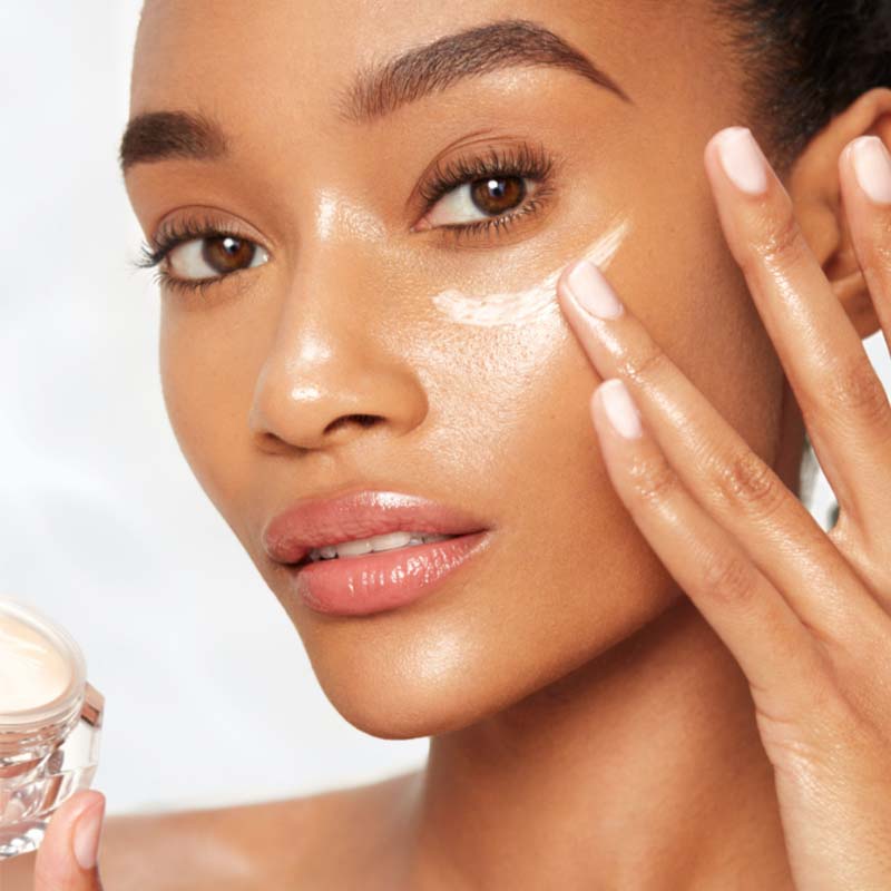 Deep-tone brunette model with glass skin applying a fawn-coloured eye cream from a petite glass jar. 
