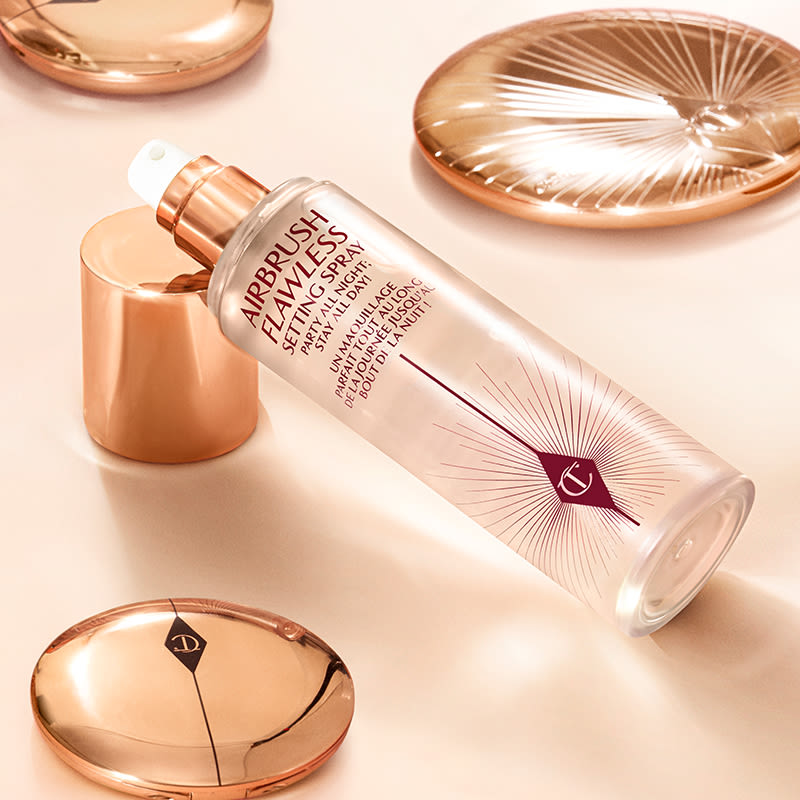 A setting spray in a clear bottle with a gold-coloured lid placed on a champagne-coloured floor with closed face powder and bronzer compacts all around.