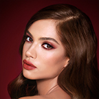 A medium-tone model with brown eyes wearing reddish-plum eye makeup, glowy face base, and a vampy-red lipstick with a satin-finish. 