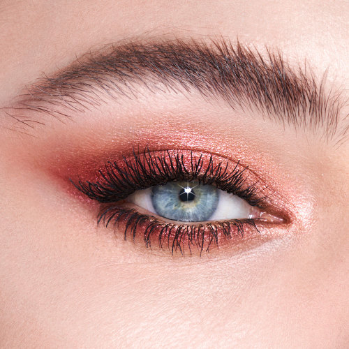 Single-eye close-up of a model with blue eyes wearing rose gold, red, and cream eye makeup.