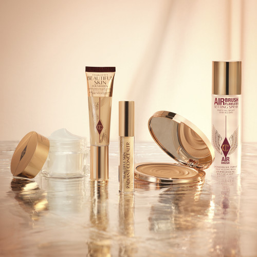 Foundation in a sleek gold-coloured tube with a concealer in a glass tube and gold-coloured lid, dark brown cream bronzer compact in gold-coloured packaging, setting spray in a large, clear bottle with a gold-coloured lid, and pearly-white face cream in a glass jar with a gold-coloured lid.
