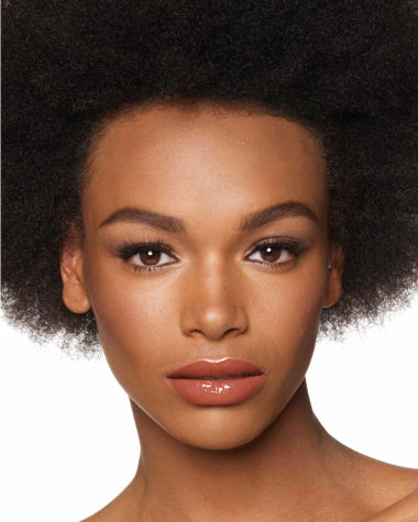 Deep-tone model with brown eyes wearing a moisturising lipstick balm in a peachy-nude shade with a high-shine finish.