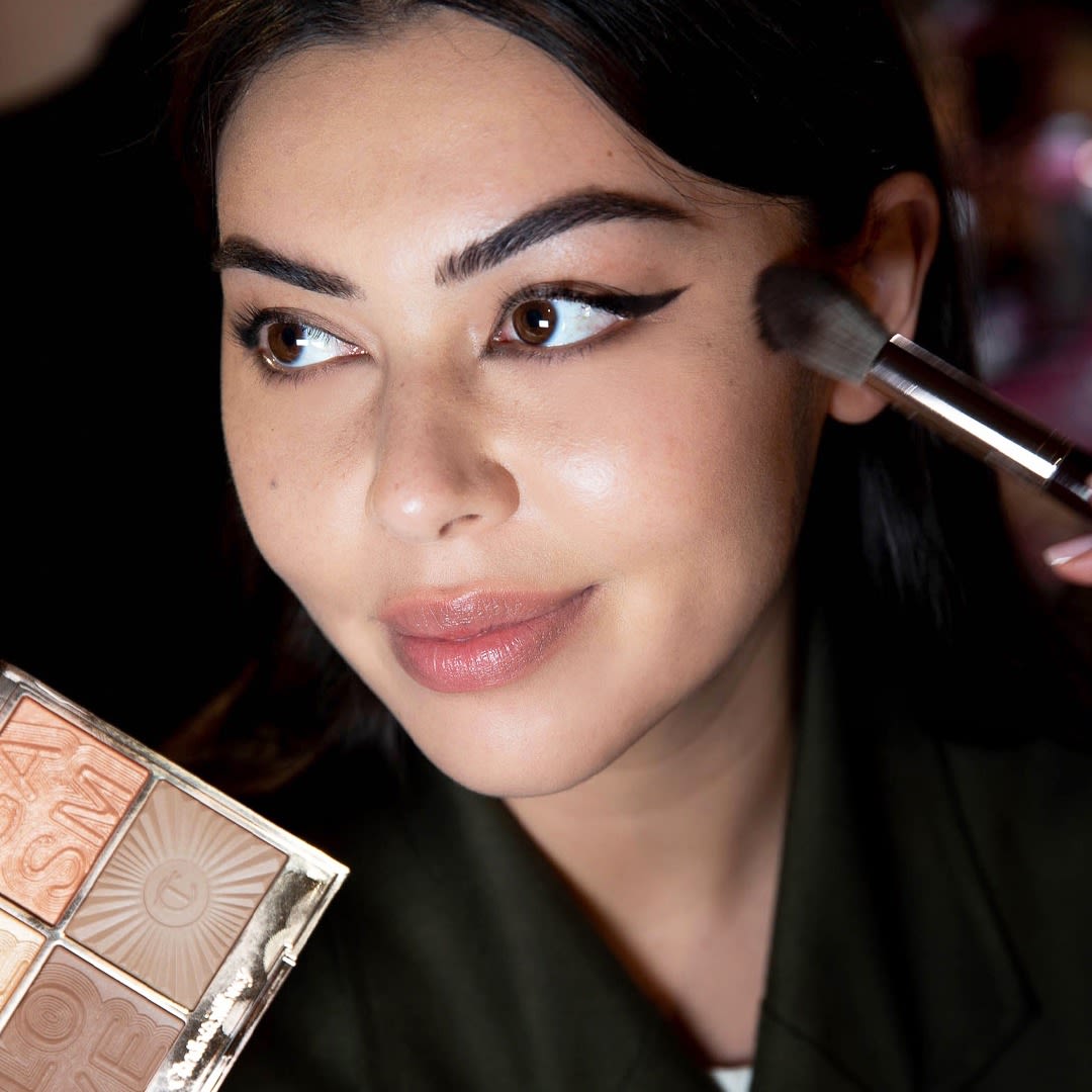 Light-tone brunette model wearing nude pink lipstick with black eyeliner and getting a glowy, peach highlighter blush applied.