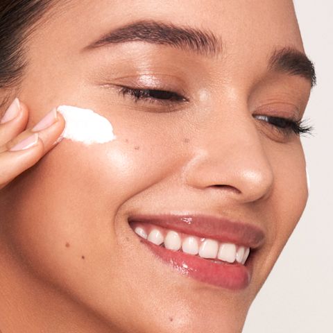 Deep-tone brunette model with glowy, flawless skin applying white-coloured face cream from a glass jar. 