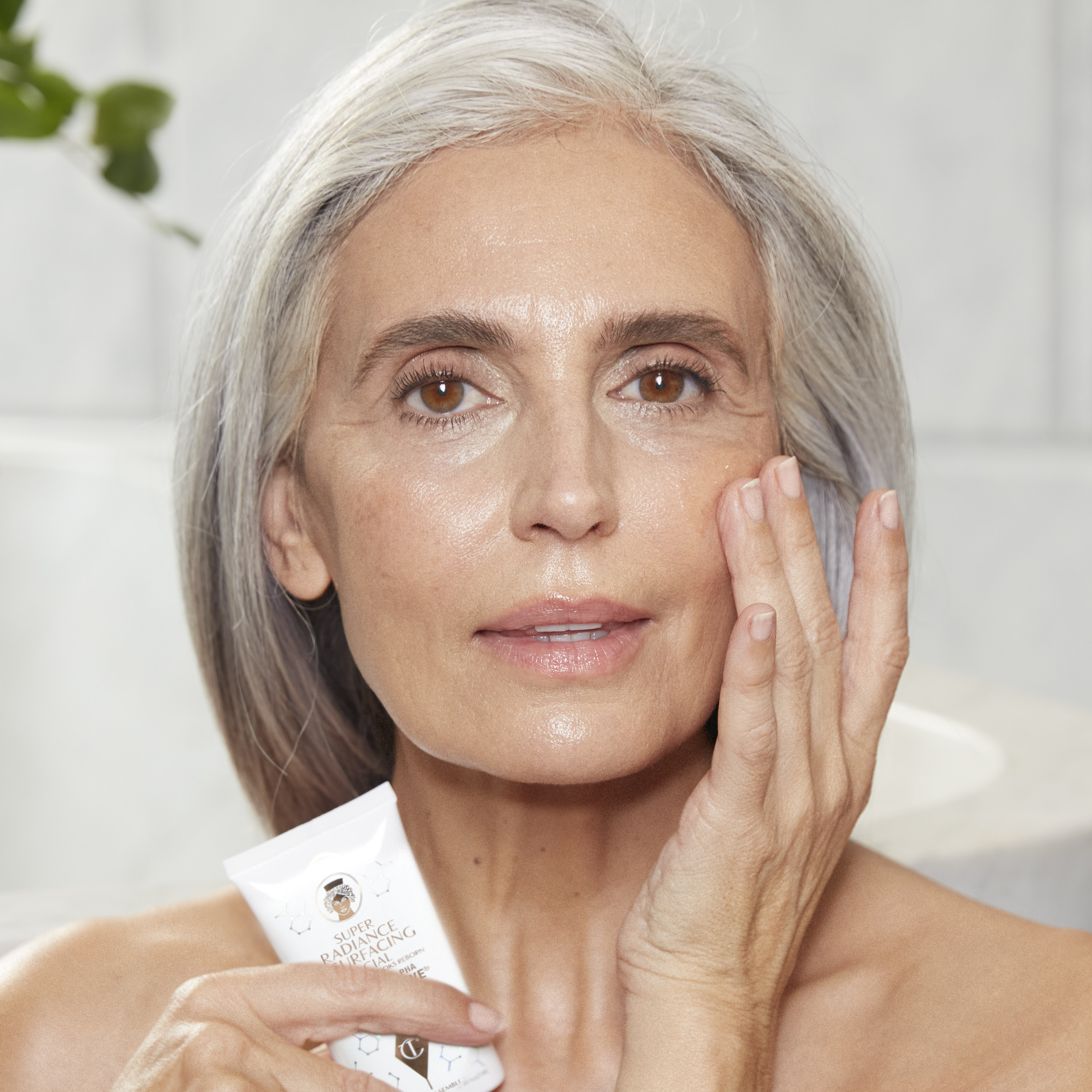 Light-tone model with brown eyes and mature, smooth, flawless, and glowy glass skin holding a white-coloured tube of a wash-off exfoliating mask.
