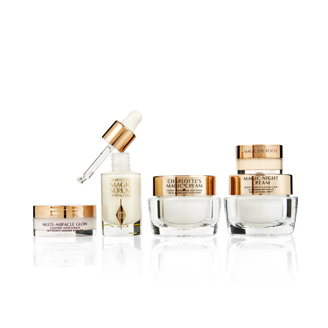 Banner with a cleansing balm in a glass jar, facial serum in a glass bottle and its dropper lid next to it, eye cream, day, and night creams in glass jars with gold-coloured lids. 