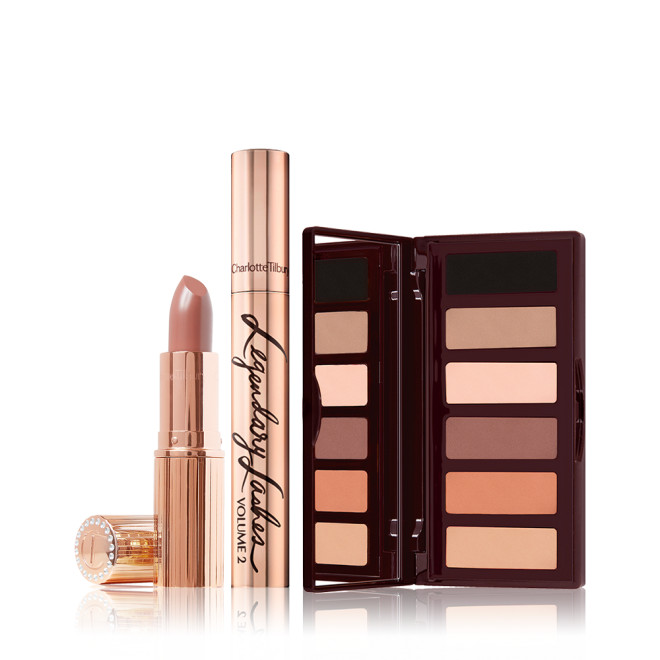 An open cool-beige lipstick with its lid next to it with a closed gold-colour mascara tube, and an open, mirrored-lid eyeshadow palette in nude brown, peach, and beige shades. 