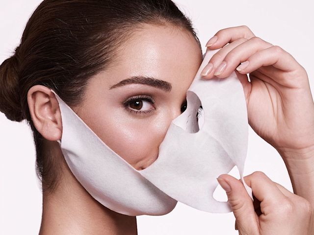 Close-up of a light-tone, brunette model easily taking off a sheet mask made of textile fabric with ear hooks and eyes, lips, and nose openings.