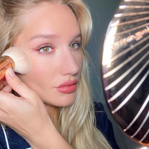 Sofia Tilbury applying bronzer while wearing soft fawn eye makeup with bright pink eyeliner and golden peachy-pink matte lipstick.