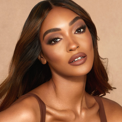 A deep-tone model with bronzed skin wearing a deep, sultry rose-brown nude lipstick with a matte finish and smokey grey and black eye makeup.