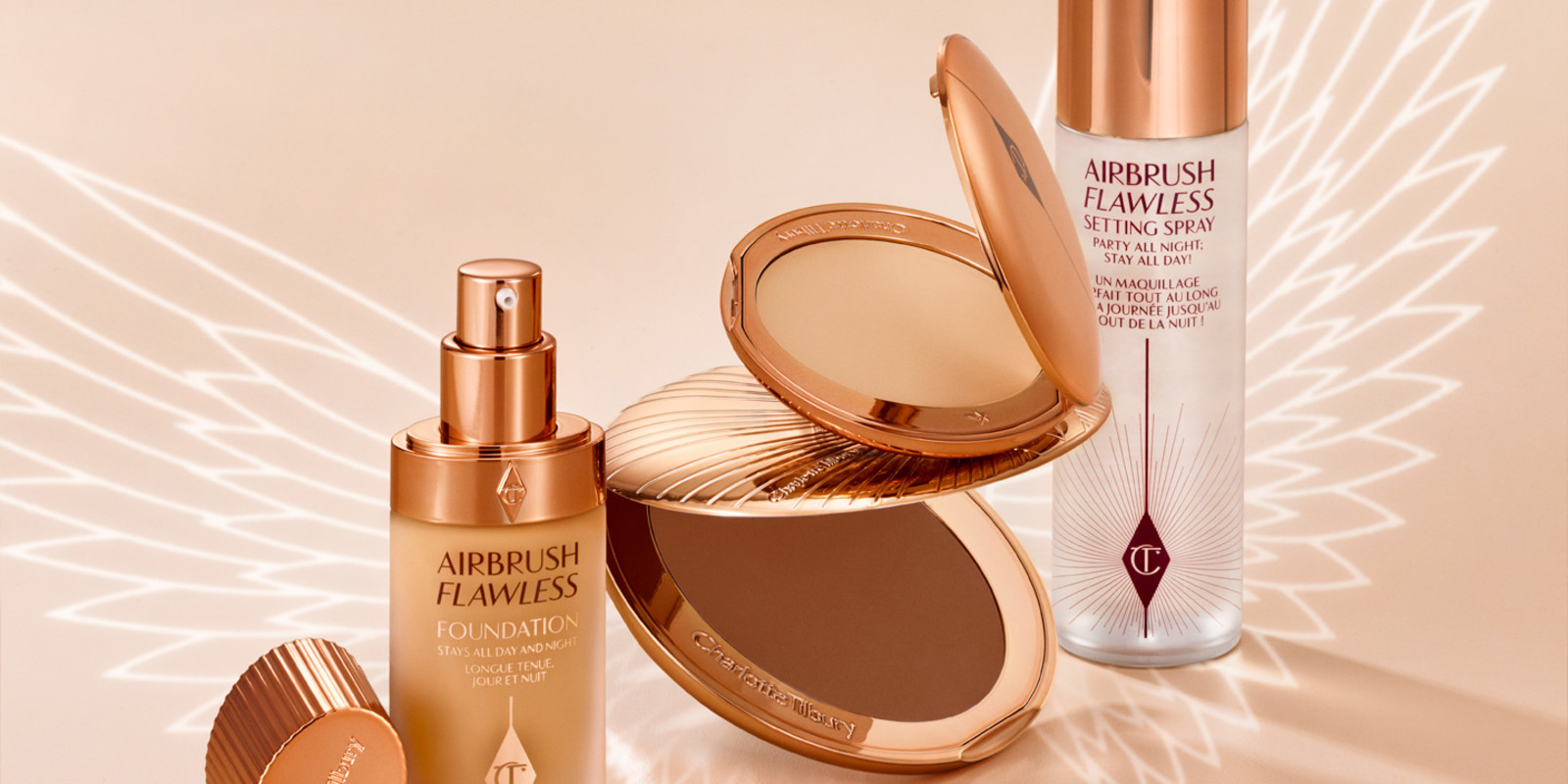 An open pressed powder compact in a light beige shade with a foundation in a frosted glass bottle with gold-coloured lid, a setting spray in a clear bottle with a gold-coloured lid, and a bronzer compact in gold-coloured packaging.