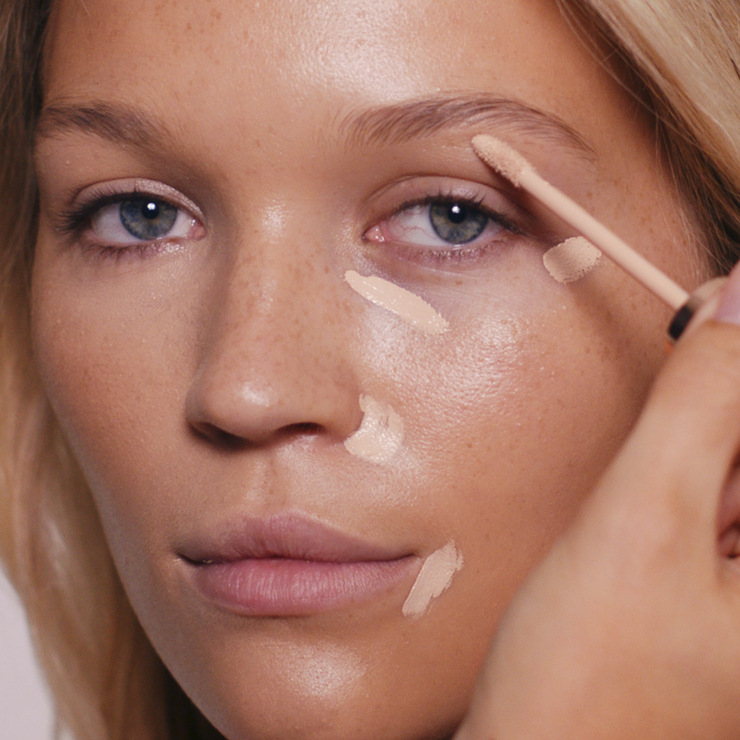 Fair model with blonde hair having concealer applied by an artist under her eyes, around the nose and around the mouth.