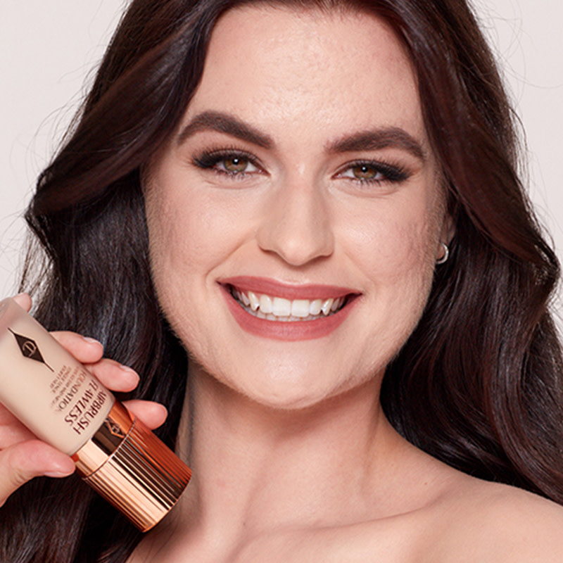 A fair-tone model with a glowy, smooth face base and wearing matte, muted red lips and nude eye makeup while holding a foundation bottle. 