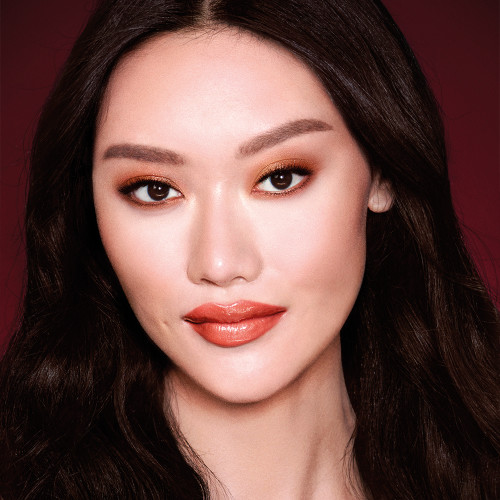 A fair-tone model with brown eyes wearing shimmery copper and gold eye makeup with black eyeliner, glowy bronzed cheeks, and pinkish-brown lipstick with gloss on top. 
