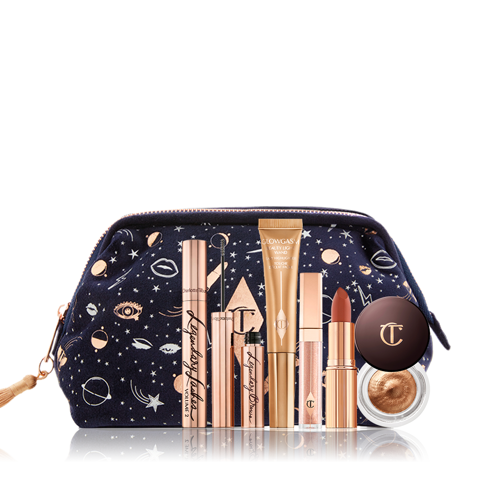 A dark blue bag with a galaxy-themed print with its makeup products displayed in front of it, mascara in a gold tube, eyebrow tint in gold tube, liquid highlighter wand, nude pink lip gloss, matte lipstick, and cream eyeshadow in a glass pot. 