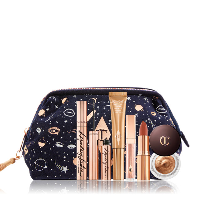 A dark blue bag with a galaxy-themed print with its makeup products displayed in front of it, mascara in a gold tube, eyebrow tint in gold tube, liquid highlighter wand, nude pink lip gloss, matte lipstick, and cream eyeshadow in a glass pot. 
