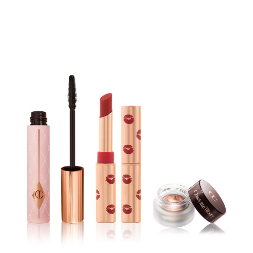 Black mascara in a sheer pink tube with a gold-coloured lid and fluffy applicator, open matte lipstick in a poppy-red colour with kiss prints printed all over the golden-coloured lipstick tube, and cream eyeshadow in an antique oyster-gold shade. in a petite pot with its lid next to it.